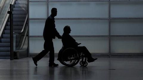 Ein junger Mann schiebt einen Rollstuhlfahrer durch eine Halle. Es könnte eine Flughafenhalle sein. Von beiden ist nur die Silhouette erkennbar. 