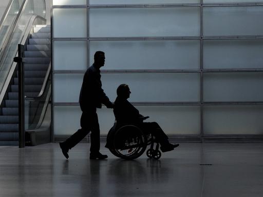 Ein junger Mann schiebt einen Rollstuhlfahrer durch eine Halle. Es könnte eine Flughafenhalle sein. Von beiden ist nur die Silhouette erkennbar. 
