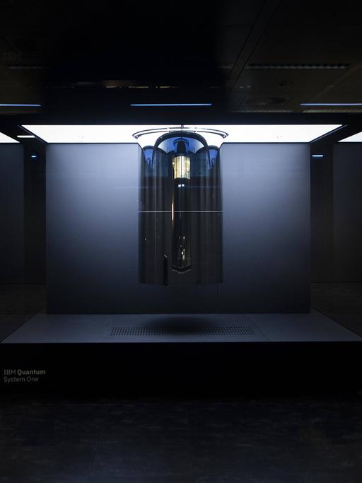 Ein futuristisch anmutendes Gebilde in einem Kasten steht in einem schwarzen Raum. Darauf steht der Name des Quantencomputer "IBM Quantum System One".