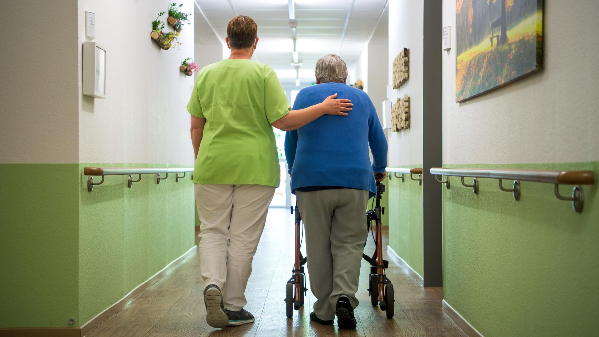 Eine Pflegefachkraft geht mit einer Bewohnerin durch den Flur eines Seniorenheims. Sie hat ihr einen Arm umgelegt. Die alte Frau geht am Rollator. 