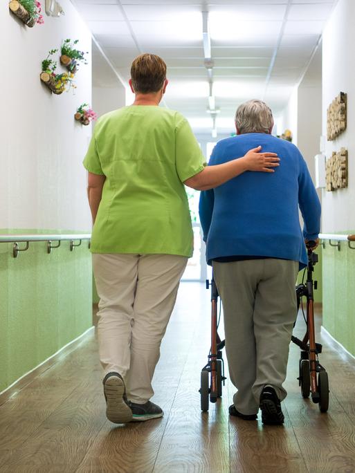 Eine Pflegefachkraft geht mit einer Bewohnerin durch den Flur eines Seniorenheims. Sie hat ihr einen Arm umgelegt. Die alte Frau geht am Rollator. 
