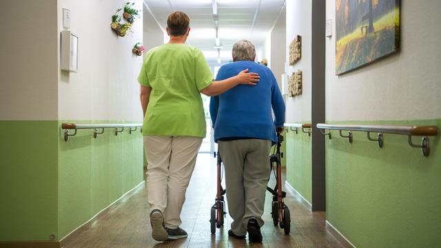 Eine Pflege-Fachkraft geht mit einer Bewohnerin durch den Flur eines Senioren-Heims.  Die alte Frau geht am Rollator. 