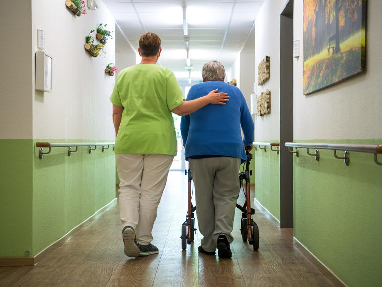 Eine Pflegefachkraft geht mit einer Bewohnerin durch den Flur eines Seniorenheims. Sie hat ihr einen Arm umgelegt. Die alte Frau geht am Rollator. Eine Pflegefachkraft geht mit einer Bewohnerin durch den Flur eines Seniorenheims. Sie hat ihr einen Arm umgelegt. Die alte Frau geht am Rollator.