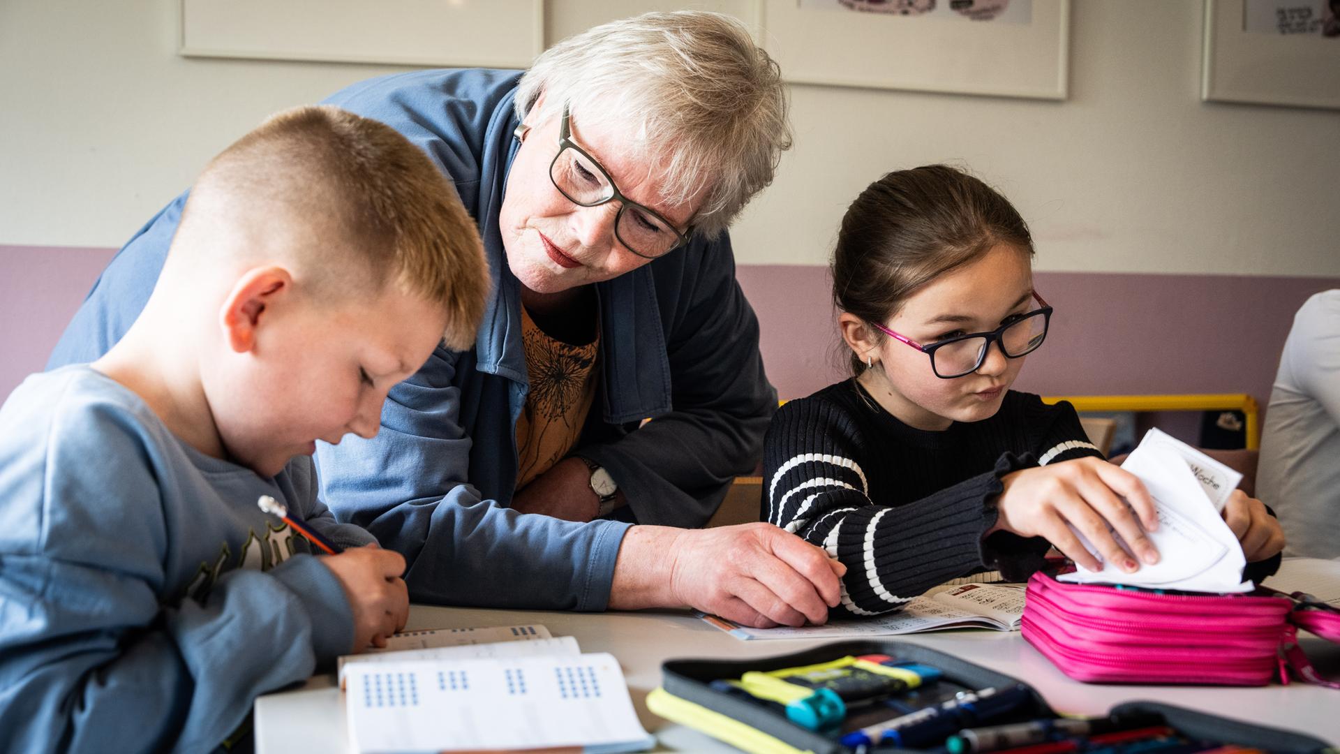 Ehrenamt in Duisburg: Eine pensionierte Lehrerin hilft zwei Kindern bei den Hausaufgaben, sie steht in der Mitte von den sitzenden Grundschulkindern, die in ihre Schulhefte schreiben.