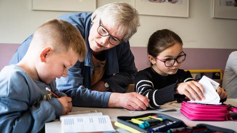 Ehrenamt in Duisburg: Eine pensionierte Lehrerin hilft zwei Kindern bei den Hausaufgaben, sie steht in der Mitte von den sitzenden Grundschulkindern, die in ihre Schulhefte schreiben. Ehrenamt in Duisburg: Eine pensionierte Lehrerin hilft zwei Kindern bei den Hausaufgaben, sie steht in der Mitte von den sitzenden Grundschulkindern, die in ihre Schulhefte schreiben.