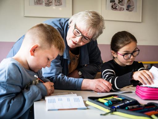 Ehrenamt in Duisburg: Eine pensionierte Lehrerin hilft zwei Kindern bei den Hausaufgaben, sie steht in der Mitte von den sitzenden Grundschulkindern, die in ihre Schulhefte schreiben.