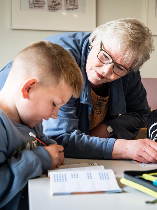 Ehrenamt in Duisburg: Eine pensionierte Lehrerin hilft zwei Kindern bei den Hausaufgaben, sie steht in der Mitte von den sitzenden Grundschulkindern, die in ihre Schulhefte schreiben.