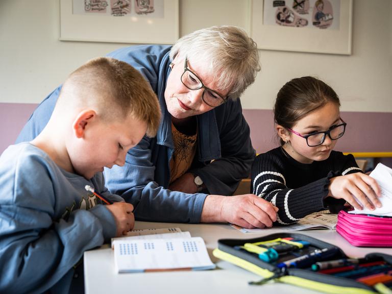 Ehrenamt in Duisburg: Eine pensionierte Lehrerin hilft zwei Kindern bei den Hausaufgaben, sie steht in der Mitte von den sitzenden Grundschulkindern, die in ihre Schulhefte schreiben.