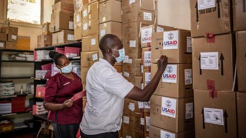 Zwei Personen beschriften in einem Lager aufeinandergestapelte Kartons mit der Aufschrift "USAID". Zwei Personen beschriften in einem Lager aufeinandergestapelte Kartons mit der Aufschrift "USAID".