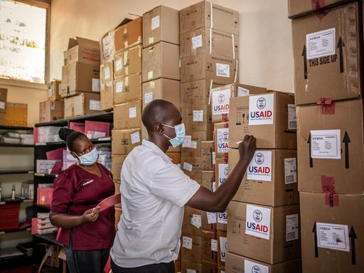 Zwei Personen beschriften in einem Lager aufeinandergestapelte Kartons mit der Aufschrift "USAID".