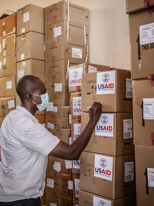 Zwei Personen beschriften in einem Lager aufeinandergestapelte Kartons mit der Aufschrift "USAID".