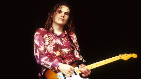 Billy Corgan von Smashing Pumpkins steht in den 90er-Jahren mit seiner Gitarre, rot geblümten Hemd und offenem Haaren auf einer Bühne.