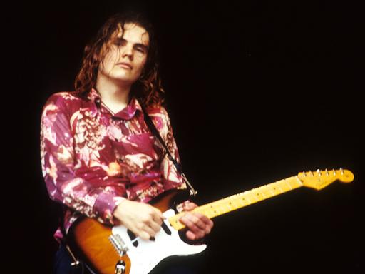 Billy Corgan von Smashing Pumpkins steht in den 90er-Jahren mit seiner Gitarre, rot geblümten Hemd und offenem Haaren auf einer Bühne.