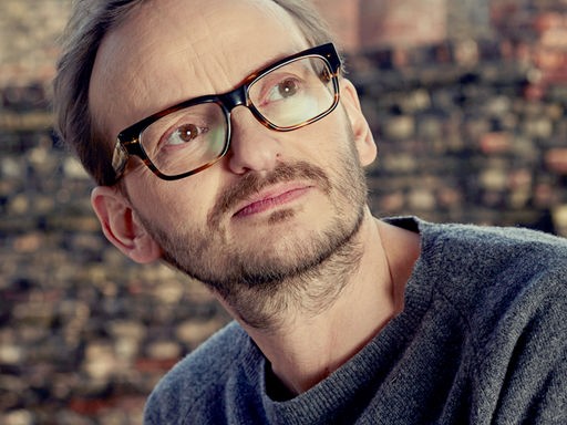 Milan Peschel steht vor einer Backsteinmauer und schaut nach rechts oben.