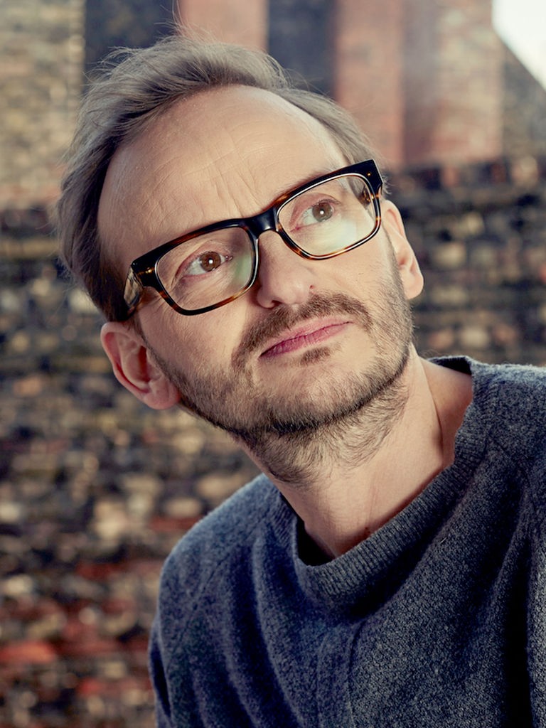 Milan Peschel steht vor einer Backsteinmauer und schaut nach rechts oben.