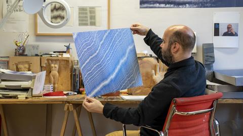 Julius Bockelt sitzt in seinem Atelier am Arbeitstisch und hält eines seiner Bilder in den Händen. Es zeigt hunderte blaue, sehr fein und eng beieinader gezeichnete Linien.
