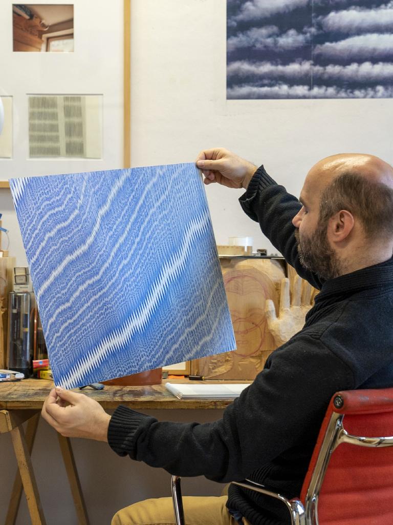 Julius Bockelt sitzt in seinem Atelier am Arbeitstisch und hält eines seiner Bilder in den Händen. Es zeigt hunderte blaue, sehr fein und eng beieinader gezeichnete Linien.