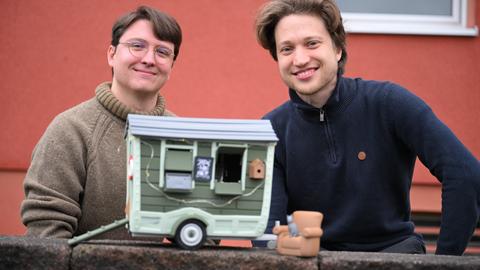 David Zapfe-Wildemann und Raven Rusch von dem Kölner Independent-Gamingstudio Neoludic stehen vor einem Haus, in dem ihre Firma ihren Sitz hat. Vor sich halten sie das Model eines Wohnwagen in Form eines Hauses. 