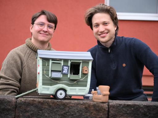 David Zapfe-Wildemann und Raven Rusch von dem Kölner Independent-Gamingstudio Neoludic stehen vor einem Haus, in dem ihre Firma ihren Sitz hat. Vor sich halten sie das Model eines Wohnwagen in Form eines Hauses. David Zapfe-Wildemann und Raven Rusch von dem Kölner Independent-Gamingstudio Neoludic stehen vor einem Haus, in dem ihre Firma ihren Sitz hat. Vor sich halten sie das Model eines Wohnwagen in Form eines Hauses.