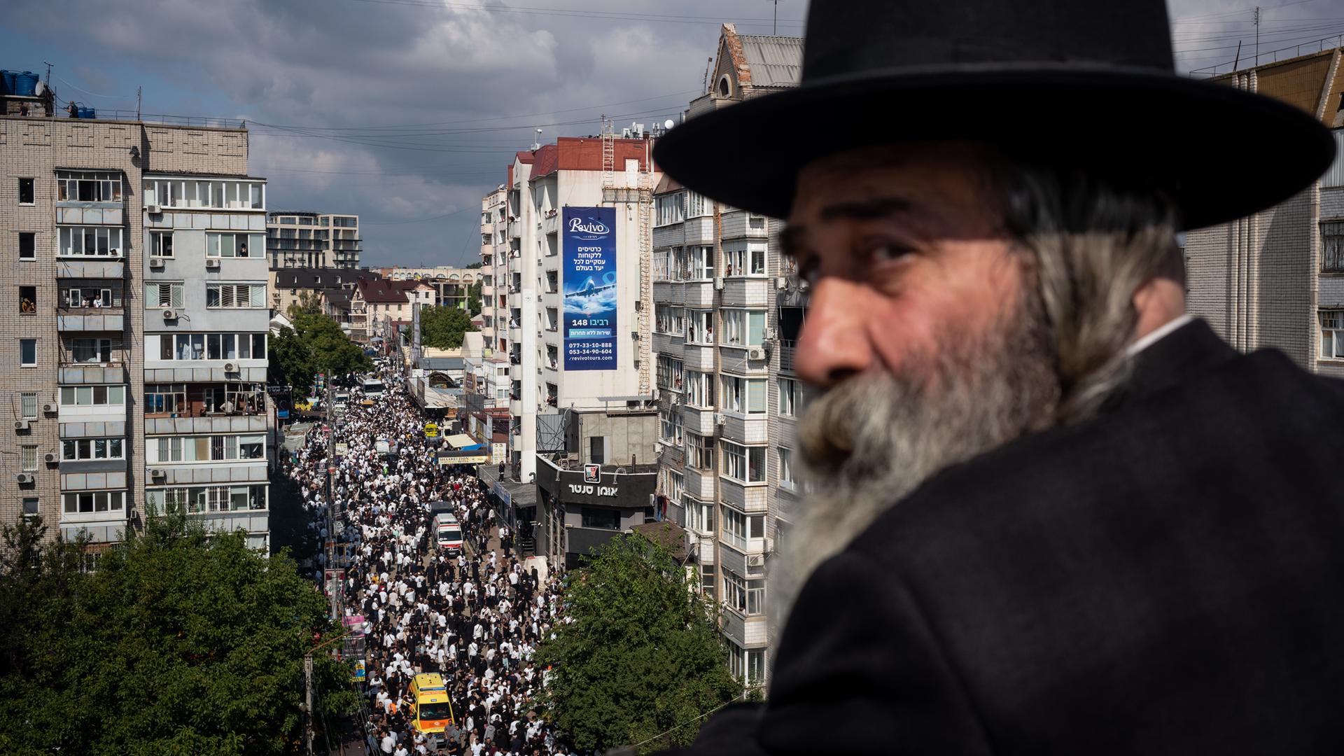 Ein alter chassidischer Jude mit langem Bart ist im Profil zu sehen. Dahinter eine Straße in der ukrainischen Stadt Uman, durch die viele Juden ziehen.