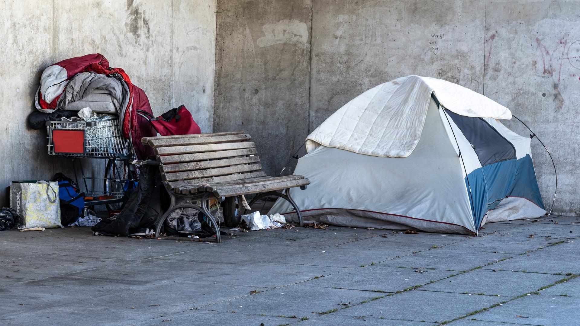 Ein Obdachlosenlager in Berlin, direkt neben dem Regierungsviertel. Ein Zelt steht neben einer Parkbank, dahinter liegen ein roter Schlafsack und Habseligkeiten eines Menschen in einem Einkaufswagen. Ein Obdachlosenlager in Berlin, direkt neben dem Regierungsviertel. Ein Zelt steht neben einer Parkbank, dahinter liegen ein roter Schlafsack und Habseligkeiten eines Menschen in einem Einkaufswagen.