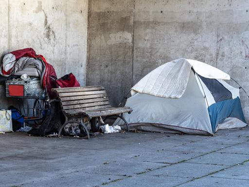 Ein Obdachlosenlager in Berlin, direkt neben dem Regierungsviertel. Ein Zelt steht neben einer Parkbank, dahinter liegen ein roter Schlafsack und Habseligkeiten eines Menschen in einem Einkaufswagen.