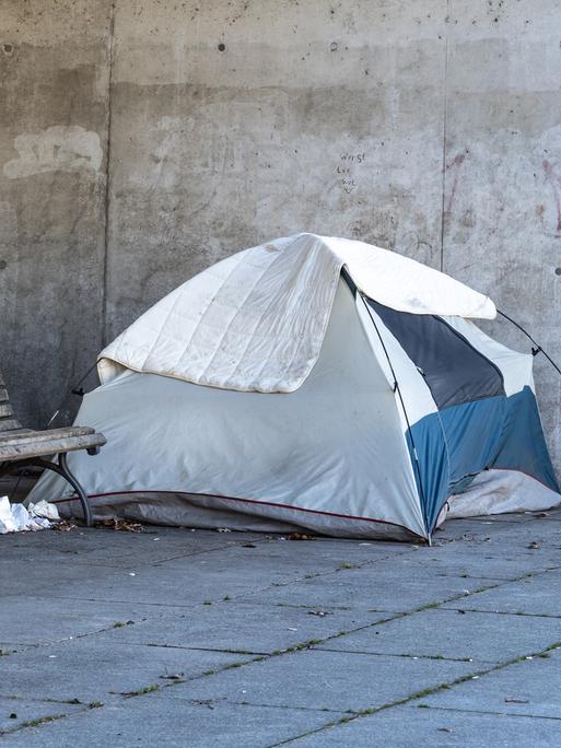 Ein Obdachlosenlager in Berlin, direkt neben dem Regierungsviertel. Ein Zelt steht neben einer Parkbank, dahinter liegen ein roter Schlafsack und Habseligkeiten eines Menschen in einem Einkaufswagen.