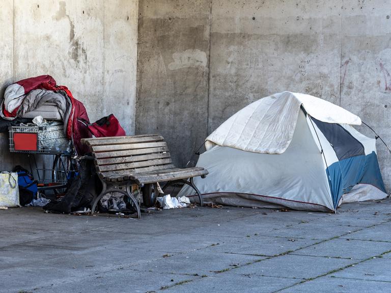 Ein Obdachlosenlager in Berlin, direkt neben dem Regierungsviertel. Ein Zelt steht neben einer Parkbank, dahinter liegen ein roter Schlafsack und Habseligkeiten eines Menschen in einem Einkaufswagen. Ein Obdachlosenlager in Berlin, direkt neben dem Regierungsviertel. Ein Zelt steht neben einer Parkbank, dahinter liegen ein roter Schlafsack und Habseligkeiten eines Menschen in einem Einkaufswagen.