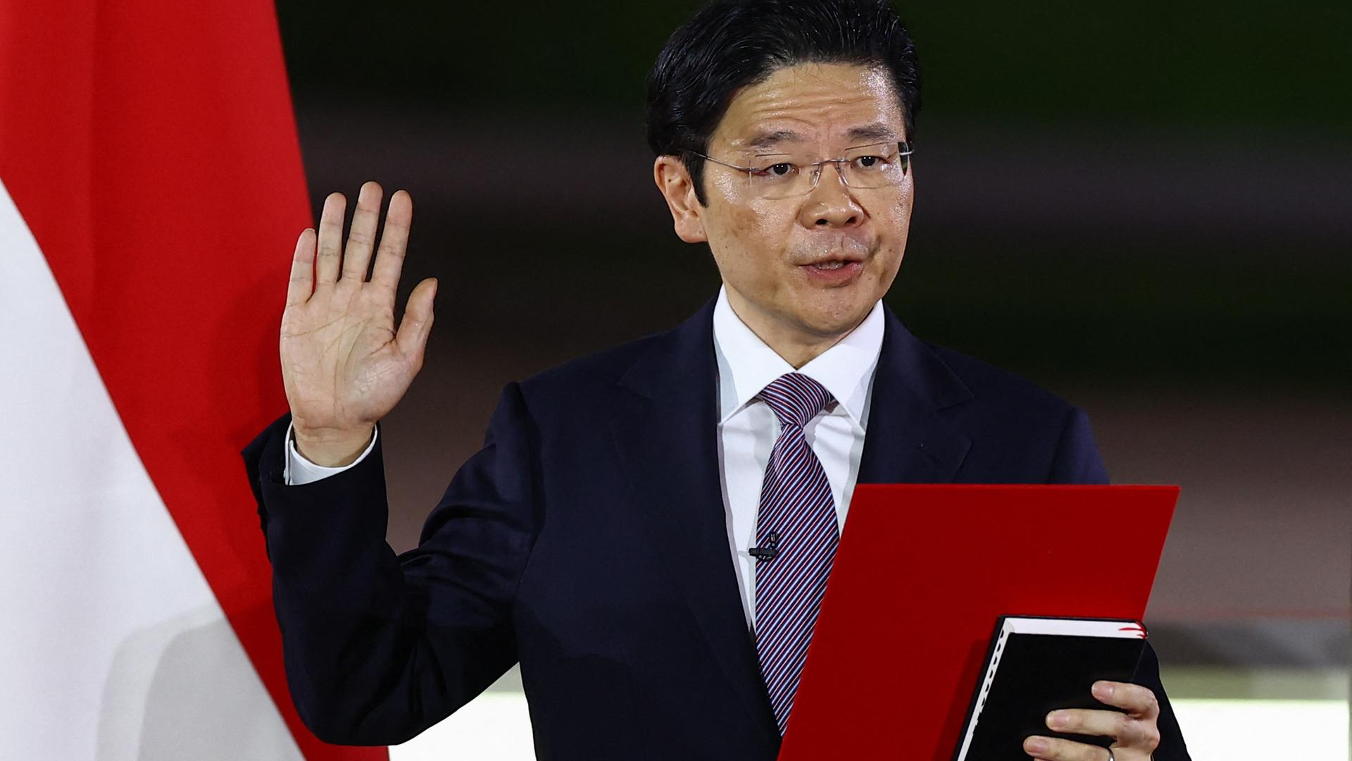 Wong im schwarzen Jackett und mit Krawatte hebt die rechte Hand und hält ein Buch - vermutlich die Bibel - sowie ein rotes Blatt in der Hand. Daneben die Flagge Singapurs.