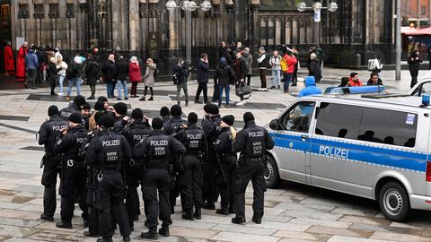24.12.2023, Nordrhein-Westfalen, Köln: Polizisten patrouillieren am Eingang des Kölner Doms am Weihnachtstag. Im Vordergrund des Bildes ist ein Mannschaftswagen der Polizei zu sehen und zahlreiche Einsatzkräfte. Im Hintergrund gehen Passanten auf den Haupteingang des Domes zu.