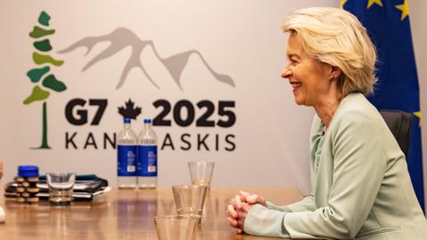 Die Präsidentin der EU-Kommission, Ursula von der Leyen, sitzt an einem Konferenztisch und lacht. Ihre Hände hat sie auf dem Tisch gefaltet. Im Hintergrund ist das G7-Logo und die EU-Flagge zu sehen. 
