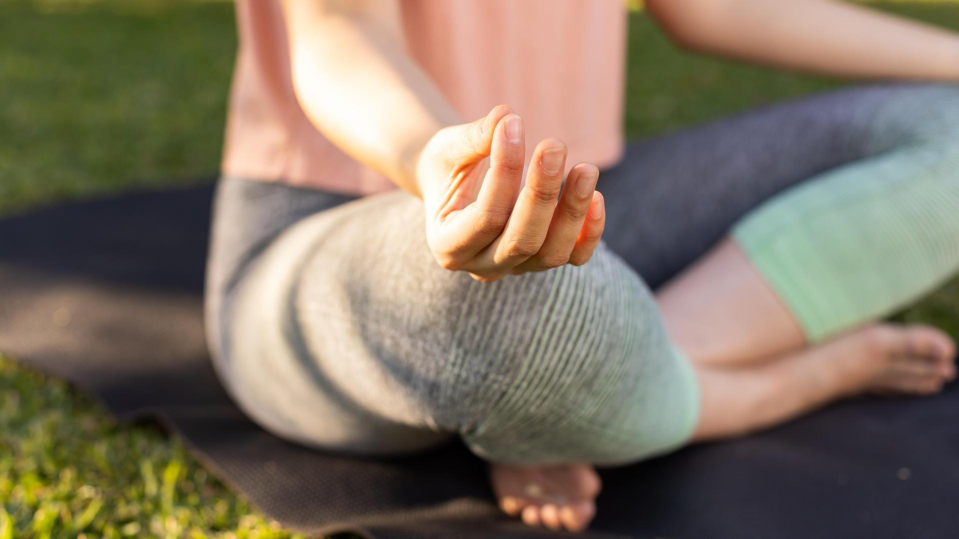Eine Frau sitzt auf einer Yogamatte und meditiert. Dabei berührt sie mit Zeige- und Mittelfinger die Kuppe des Daumens. 