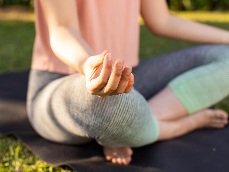 Eine Frau sitzt auf einer Yogamatte und meditiert. Dabei berührt sie mit Zeige- und Mittelfinger die Kuppe des Daumens. 
