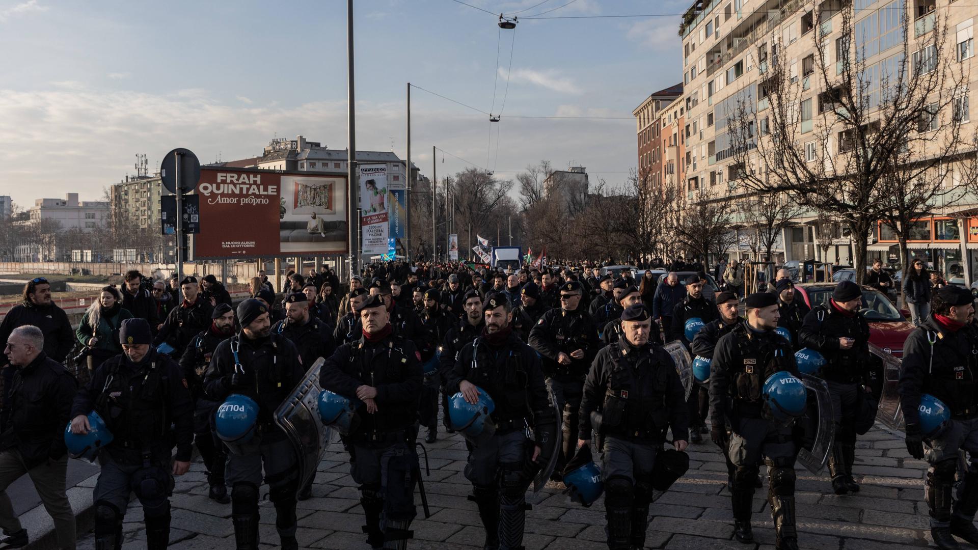 Hunderte Menschen gehen über eine Straße, vor ihnen läuft eine Reihe von Einsatzkräften mit blauen Helmen und Schutzschilden in den Händen.