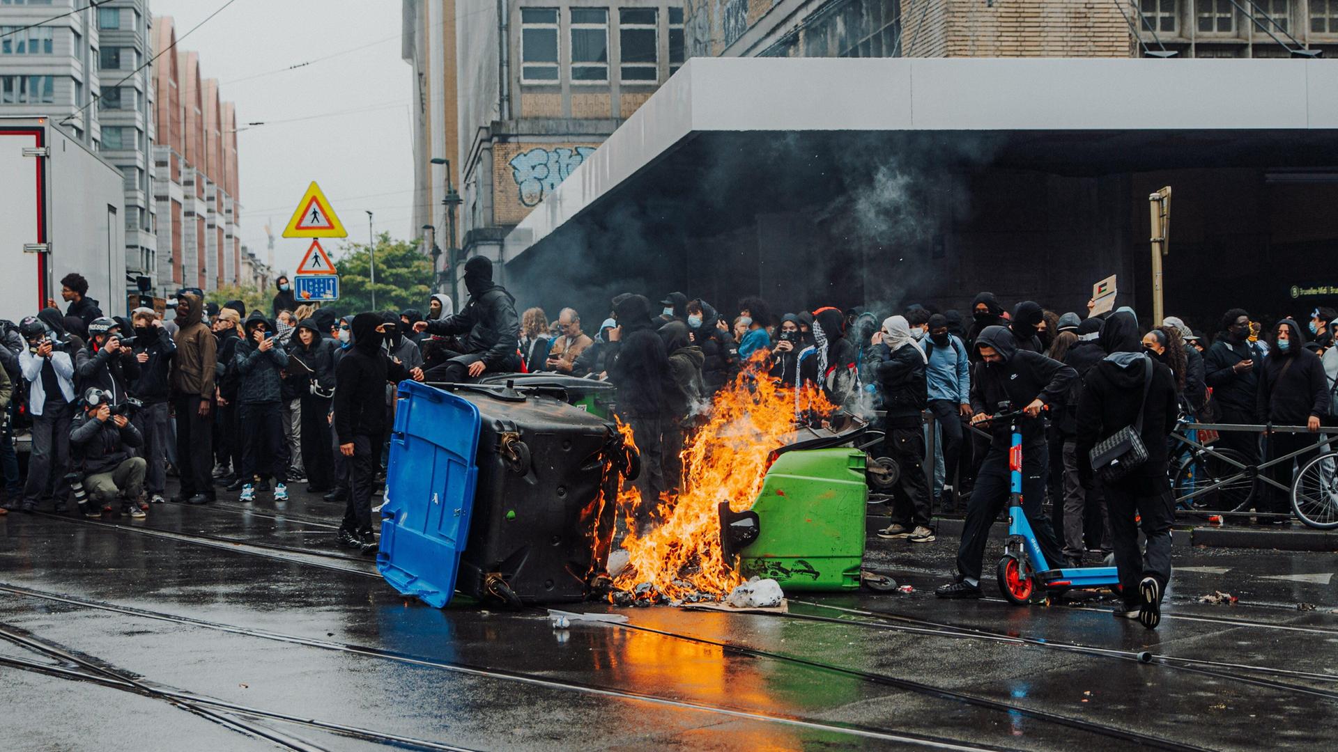 Viele schwarz gekleidete Menschen stehen in einer Gruppe, im Vordergrund brennen zwei Müllcontainer.