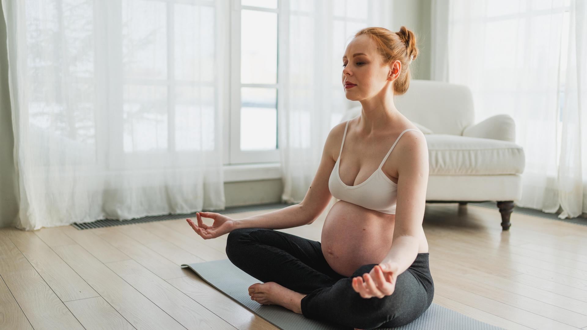 Eine hochschwangere Frau meditiert auf der Yogamatte. 