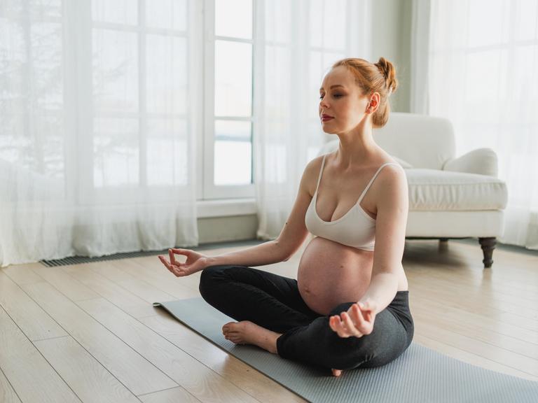 Eine hochschwangere Frau meditiert auf der Yogamatte. 
