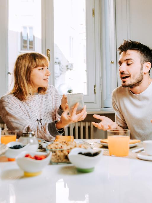 Ein Paar sitzt an einem Frühstückstisch während eine der beiden Personen ein Smartphone in der Hand hält.
