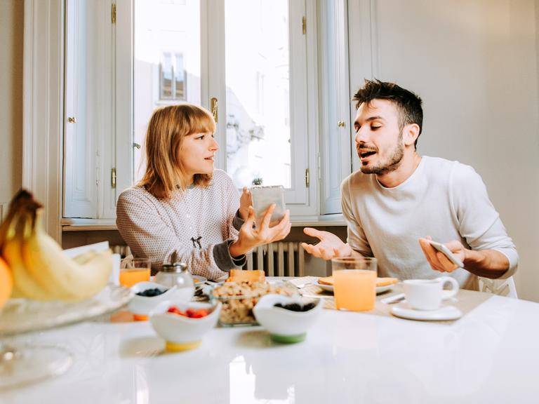 Ein Paar sitzt an einem Frühstückstisch während eine der beiden Personen ein Smartphone in der Hand hält.