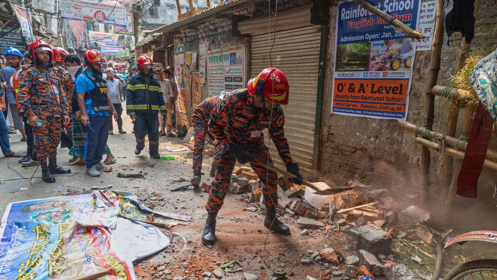 Mehrere Männer mit Helmen und Schutzanzügen stehen in einer von Schutt übersäten Gasse vor einer beschädigten Hausfassade. Mehrere Männer mit Helmen und Schutzanzügen stehen in einer von Schutt übersäten Gasse vor einer beschädigten Hausfassade.