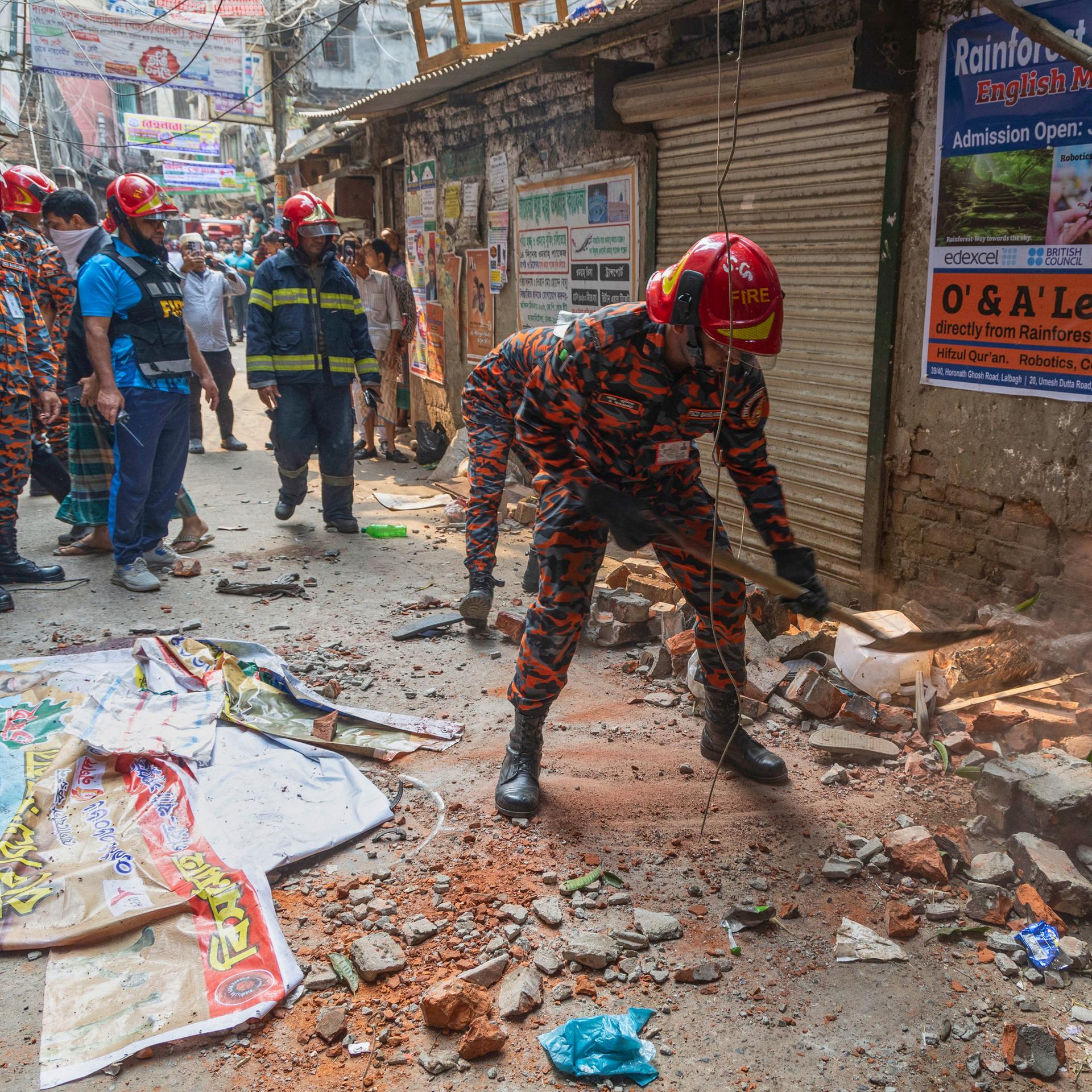 Mehrere Männer mit Helmen und Schutzanzügen stehen in einer von Schutt übersäten Gasse vor einer beschädigten Hausfassade.