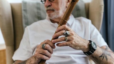 Ein älterer Mann in einem Sessel hält eine Flöte, auf die er seine Finger mit auffälligen Ringen platziert hat.