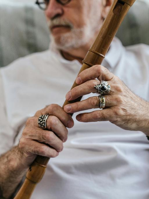 Ein älterer Mann in einem Sessel hält eine Flöte, auf die er seine Finger mit auffälligen Ringen platziert hat.