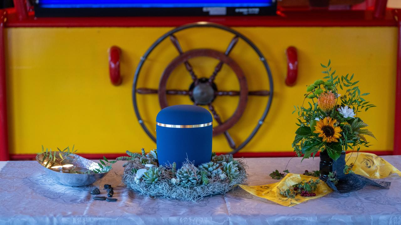 Eine geschmückte blaue Urne steht an Bord eines ehemaligen Postfrachtschiffs auf einem Podest, daneben ein Sonnenblumenstrauß.