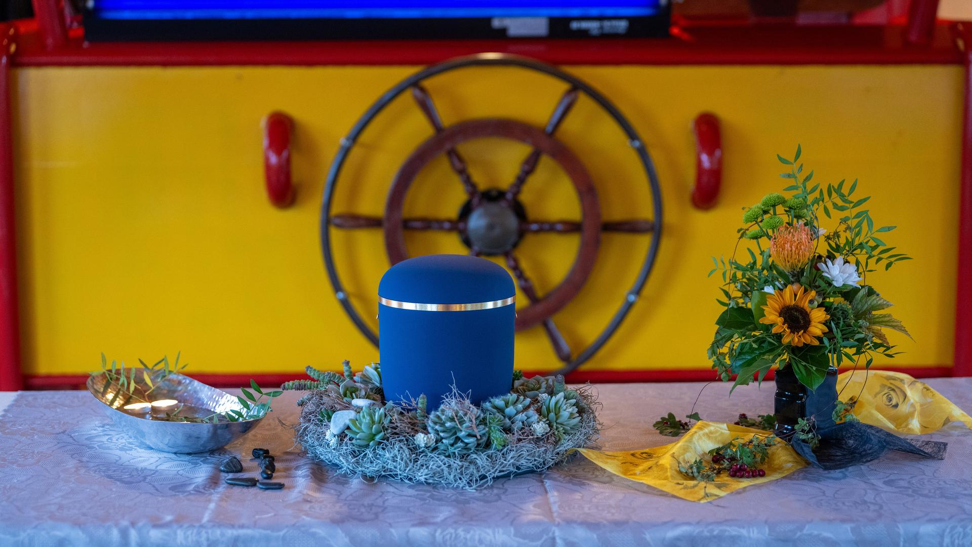 Eine geschmückte blaue Urne steht an Bord eines ehemaligen Postfrachtschiffs auf einem Podest, daneben ein Sonnenblumenstrauß.