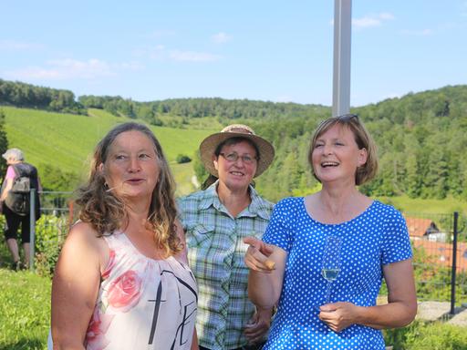 Drei Landfrauen stehen zusammen, im Hintergrund sind Felder und Wald zu erkennen.