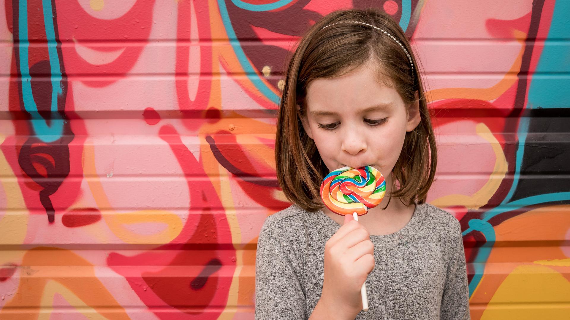 Ein Mädchen steht mit einem farbenfrohen Lolli vor einer bunten Wand