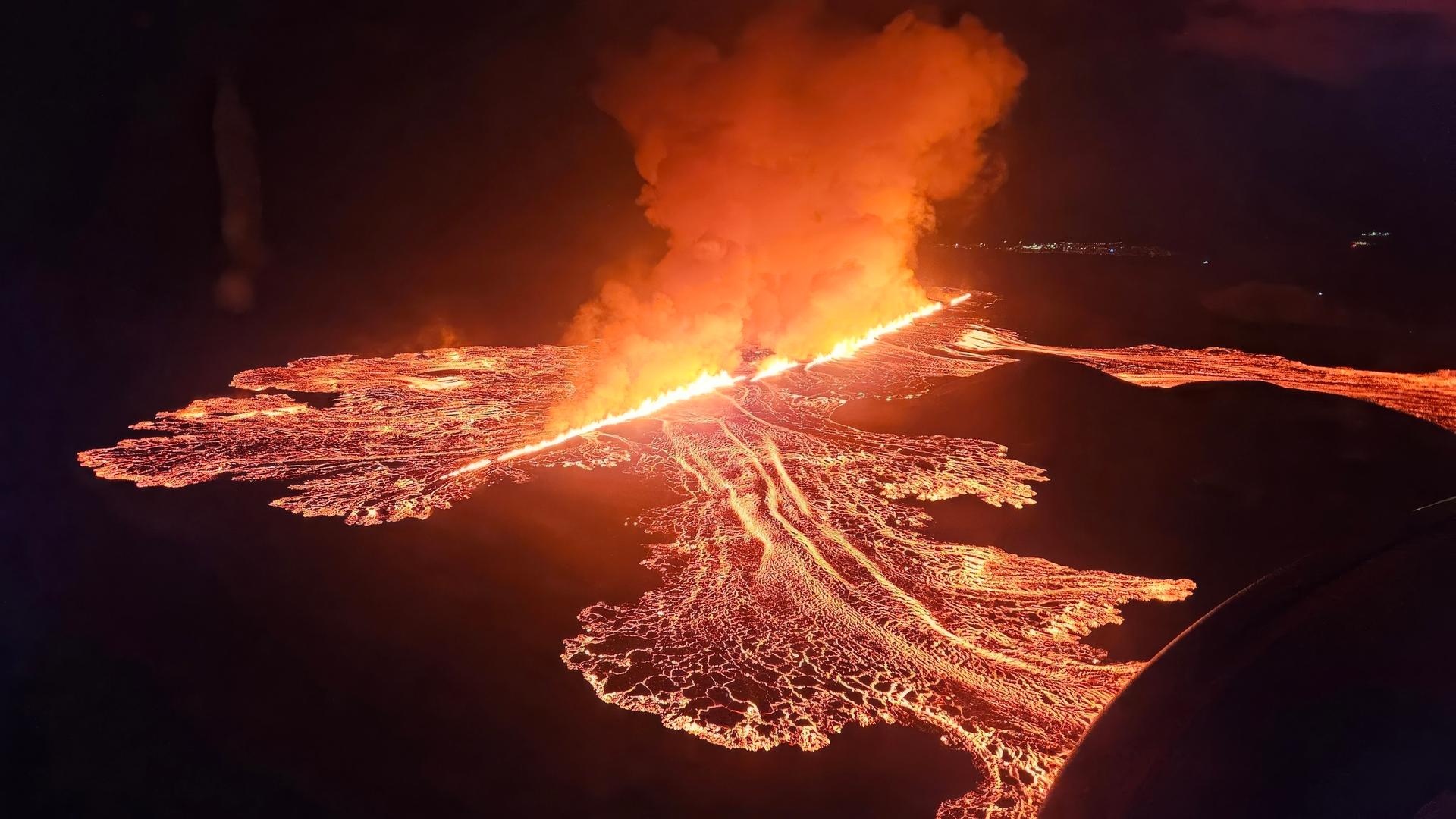 Zu sehen ist eine lange Feuerschnur, die sich durch eine dunkle Nachtlandschaft zieht. Das Feuer stammt aus einer Erdspalte vulkanischen Ursprungs. Links und rechts neben der Feuerschnur hat sich ein Geflecht aus dünneren Lava-Schnüren entsponnen. 