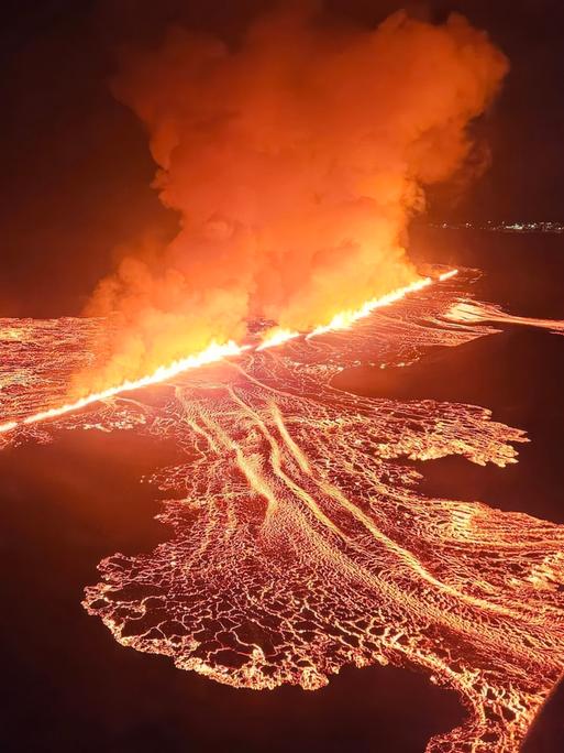 Zu sehen ist eine lange Feuerschnur, die sich durch eine dunkle Nachtlandschaft zieht. Das Feuer stammt aus einer Erdspalte vulkanischen Ursprungs. Links und rechts neben der Feuerschnur hat sich ein Geflecht aus dünneren Lava-Schnüren entsponnen. 