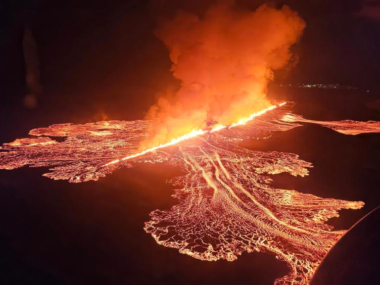 Zu sehen ist eine lange Feuerschnur, die sich durch eine dunkle Nachtlandschaft zieht. Das Feuer stammt aus einer Erdspalte vulkanischen Ursprungs. Links und rechts neben der Feuerschnur hat sich ein Geflecht aus dünneren Lava-Schnüren entsponnen. 